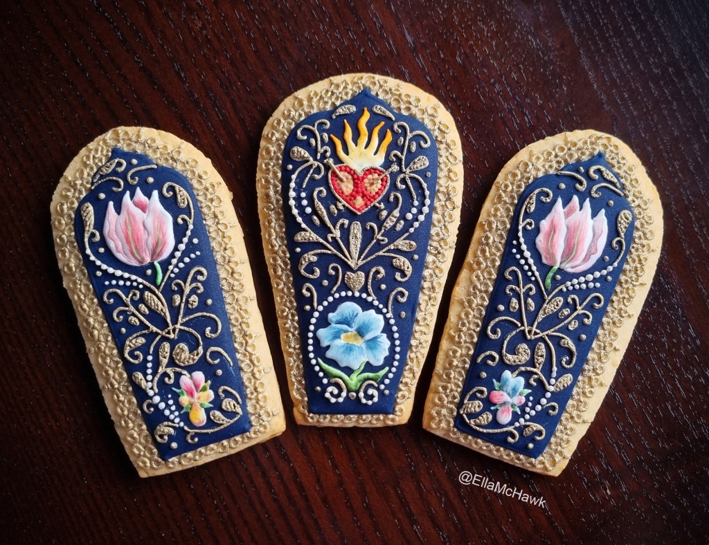 A set of three petal-shaped biscuits arranged into a fan against a dark wooden background. The biscuits are covered with a deep, rich blue icing and embellished with intricate details. At the centre of the arrangement is a textured red heart with bright yellow flames, pierced with two tiny gold arrows. The heart is surrounded by curling gold vines, delicate pearl details, and hand-painted flowers. There is a large blue pansy, two pink tulip-like flowers, and two smaller pansies in shades of pink, yellow, and blue. Each biscuit is edged with gold lace made up of small icing circles.