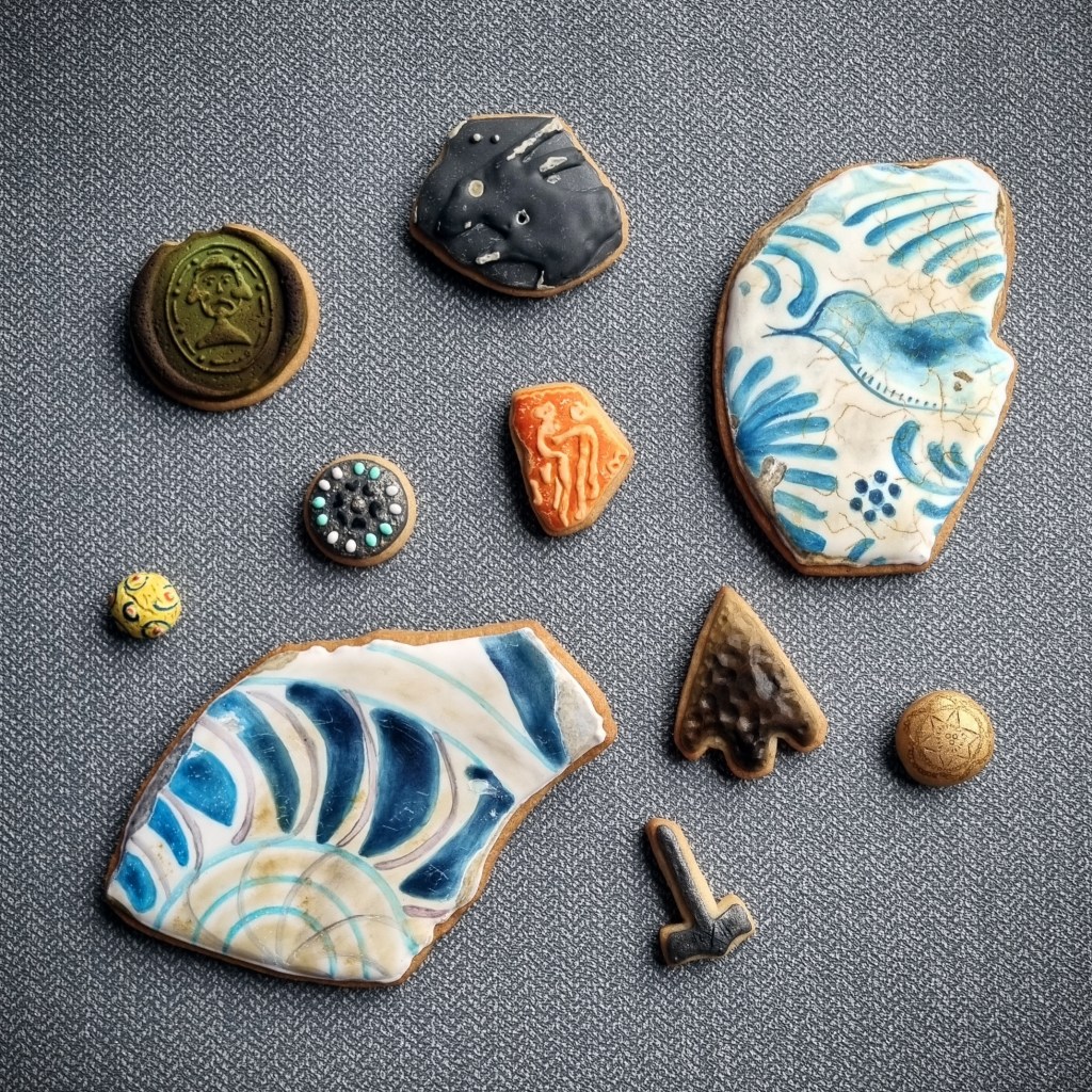 A collection of ten biscuits that resemble aged, chipped mudlarking finds. An oval biscuit in a rich, dark green looks like the seal on a medieval glass bottle, and features a raised characterful face at its centre. Next to it is an unevenly shaped black biscuit with grey chips and the raised shape of a hare’s face, ears, and upper legs. There are two large biscuits with jagged edges, each decorated in blue and white to look like broken pieces of delftware plates. One features the shape of a small bird surrounded by swirling lines, the other has a series of concentric circles and curved shapes. A small biscuit with a multifaceted texture replicates a knapped flint arrowhead. Next to it are a tiny hammer-shaped biscuit in pewter with delicate cross markings, and a very small gold biscuit button etched with lines, circles, and a rough star shape. The smallest biscuit is made to look like a yellow glass cufflink with decorative circles in blue, white, and red. Another biscuit button has the appearance of aged metal and is edged with decorate white and turquoise dots. Finally, there is a small angular biscuit in a terracotta colour with raised lines roughly in the shape of two intwined figures.